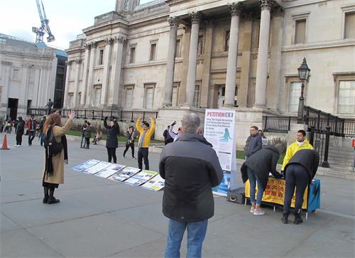 '圖4：二零一九年二月二日，法輪功學員在倫敦特拉法加廣場（Trafalgar Square）北平台的功法展示吸引過路民眾現場學煉，圖中一位西人女士正在模仿法輪功第二套功法「法輪樁法」中的「頭頂抱輪」動作。'